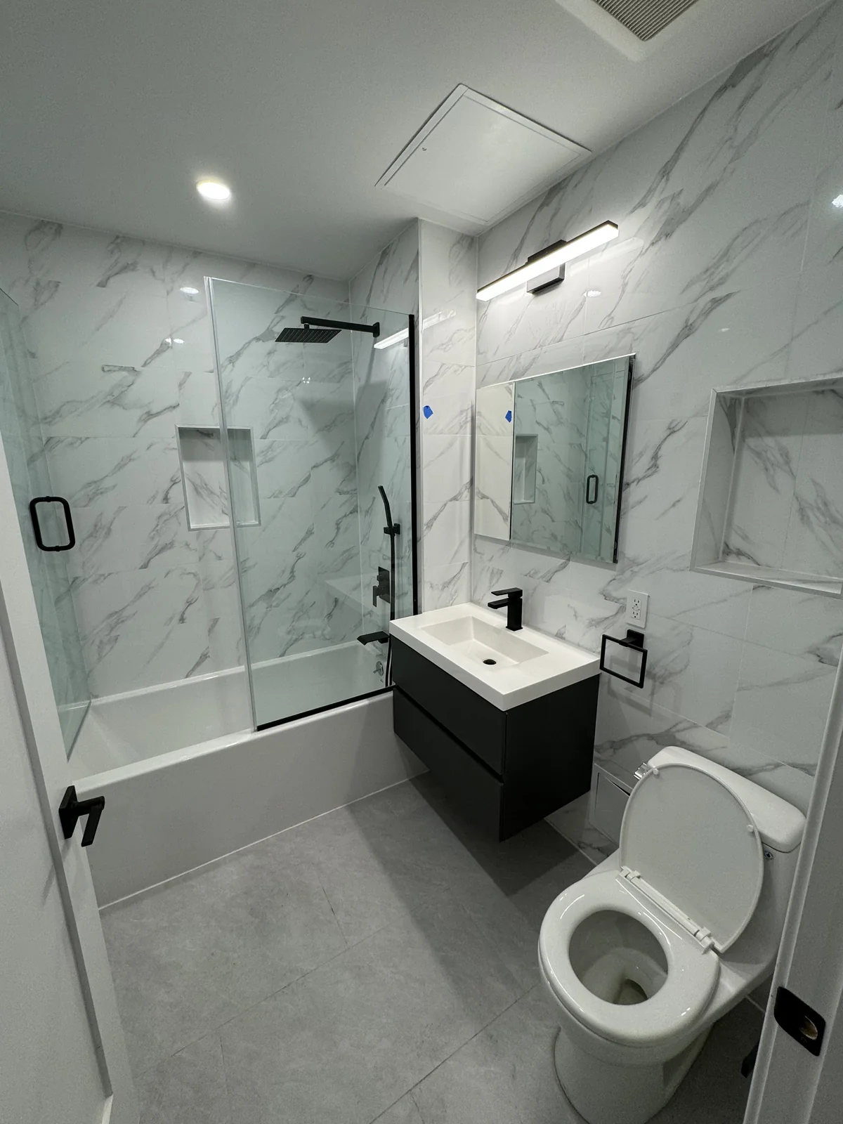 Bathroom with veined wall tile, a dark floating vanity, black fixtures, and a bathtub with a sliding glass enclosure