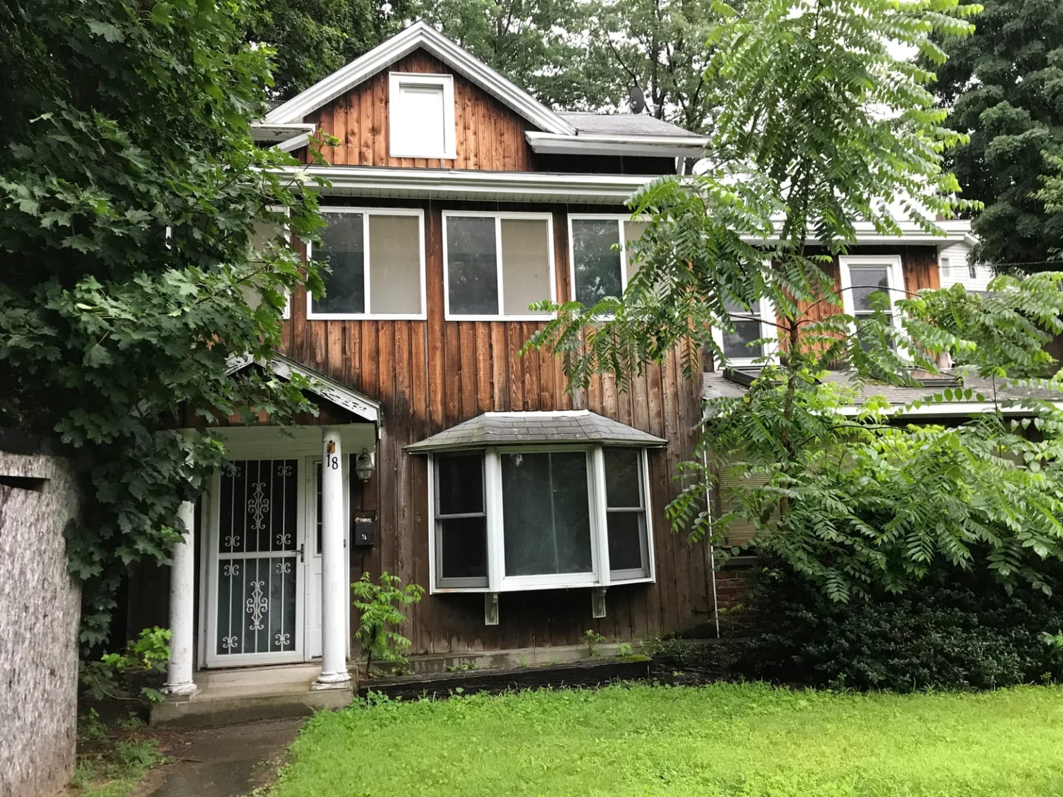 House before renovation with wood siding, white entry columns, and vegetation around the windows
